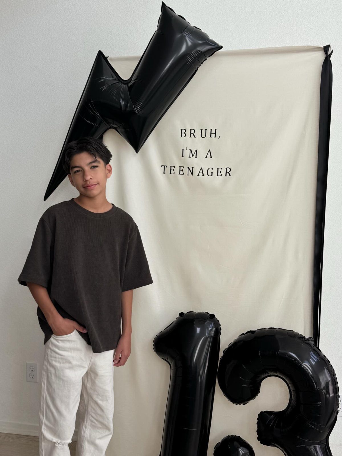 Teenage boy in brown shirt and white trousers posing by 13 balloons