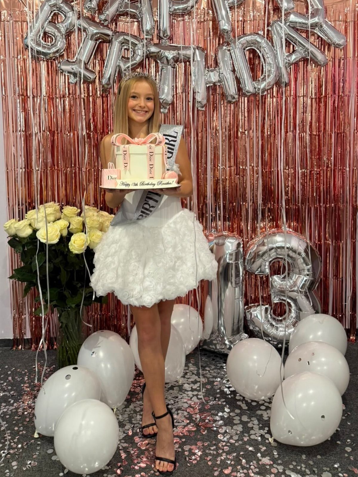 Girl in white one-shoulder fluffy dress holding cake with 13th birthday decorations