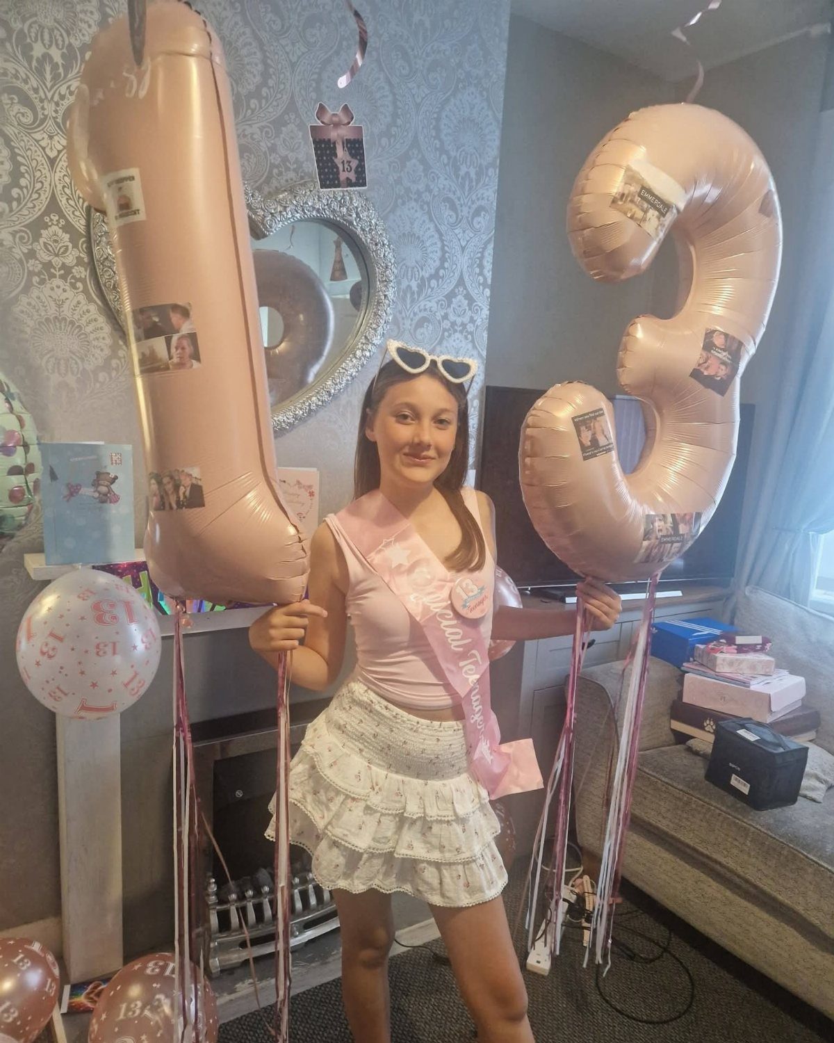 Teenage girl posing with rose-gold 13 balloons wearing a pink top and white skirt.