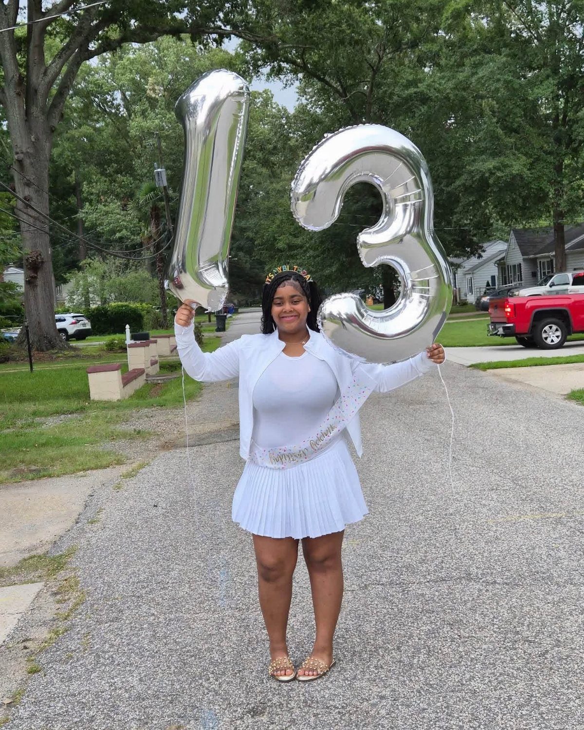 Girl in white outfit holding silver number 13 balloons on a residential street