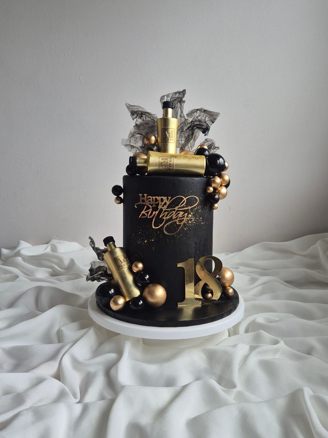 Black birthday cake with gold spheres, gold miniature bottles, and a gold 18 topper.