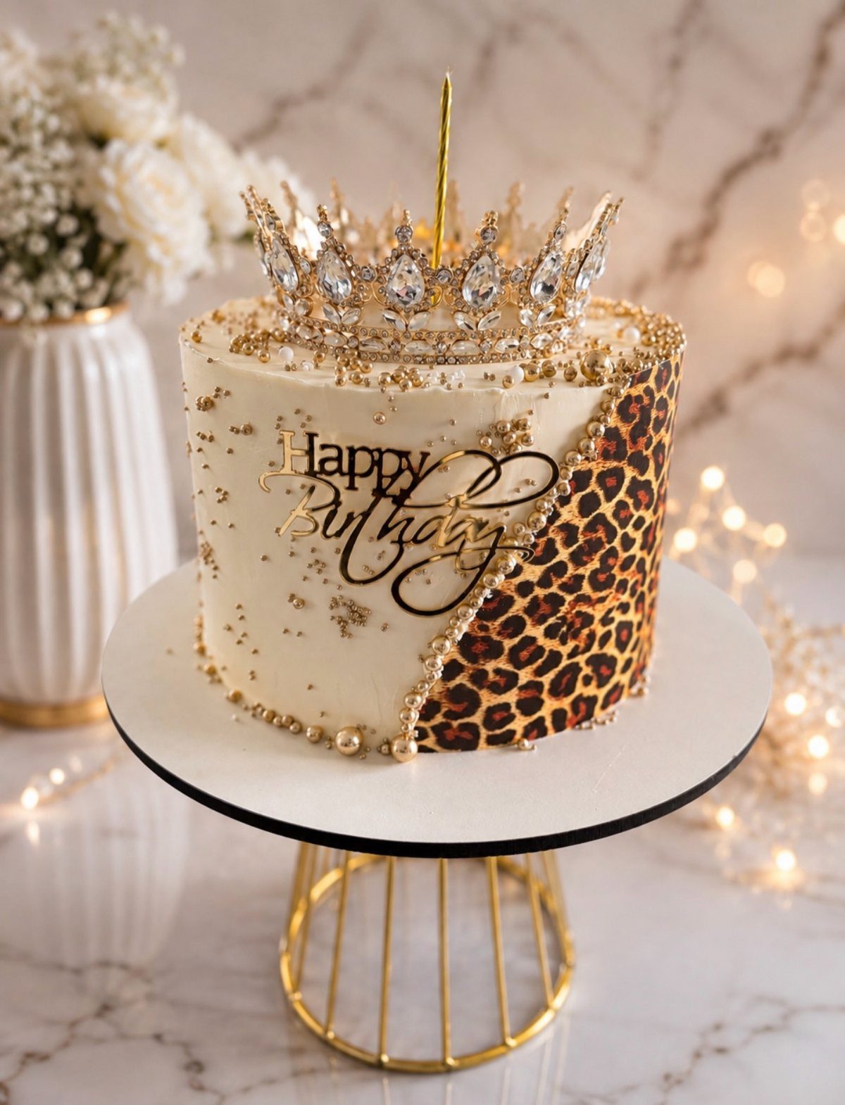 White birthday cake with leopard print, gold pearls, and a jeweled crown topper.