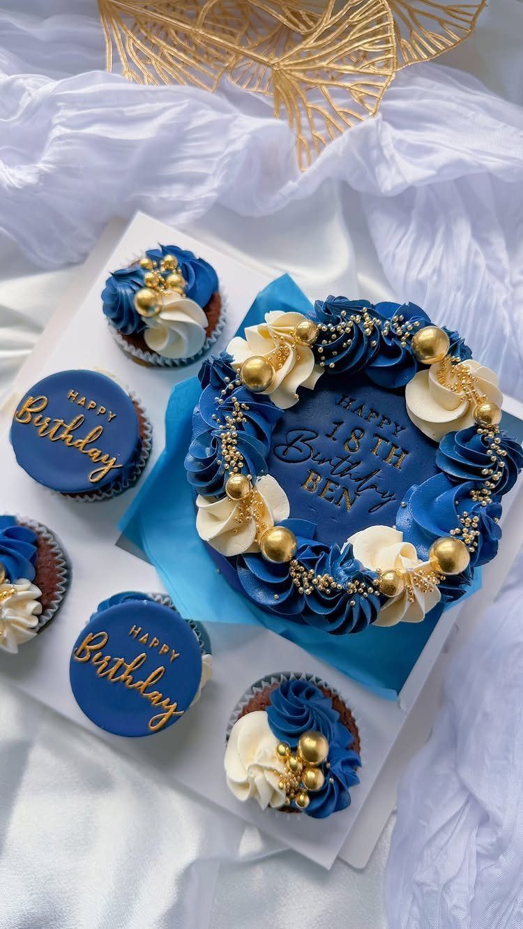 Royal blue birthday cake and cupcakes with gold pearls and white frosting swirls.
