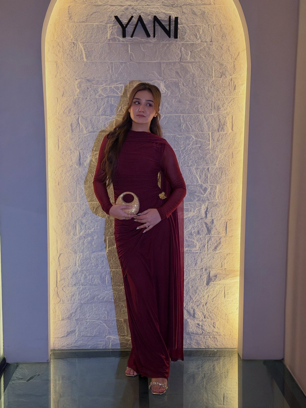 Woman in a burgundy ruched maxi dress holding a gold spherical clutch bag.