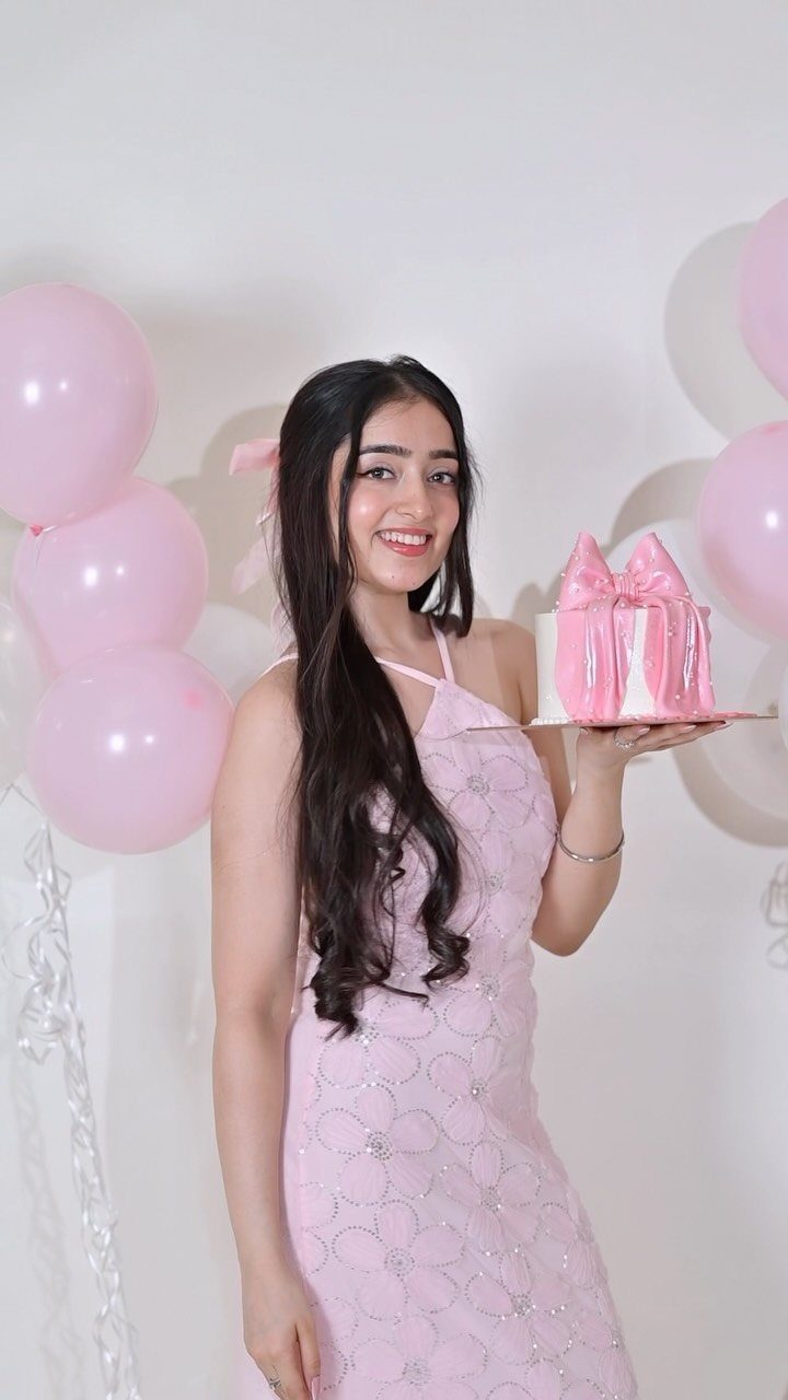 Young woman in pink dress holding a pink cake with pink balloons behind her.