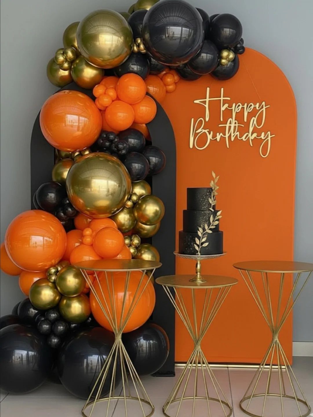 Orange and black birthday backdrop with gold balloon garland and tiered black cake.