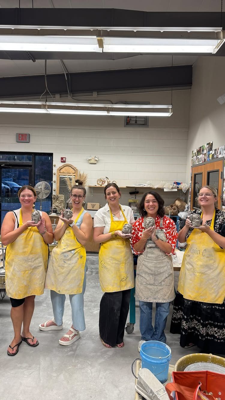 Five women in aprons holding handmade ceramic pots in a pottery studio.