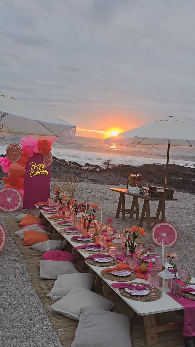 Beach birthday dinner setup with pink decorations and cushions during a sunset