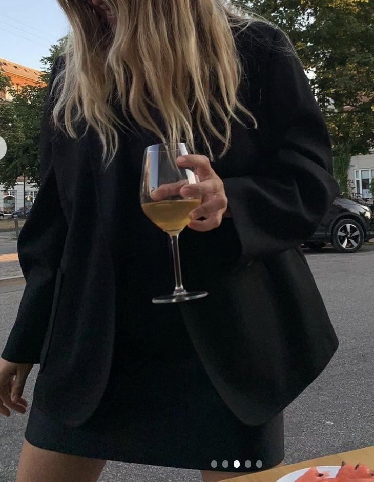 Woman in a black mini dress and oversized black blazer holding a wine glass.