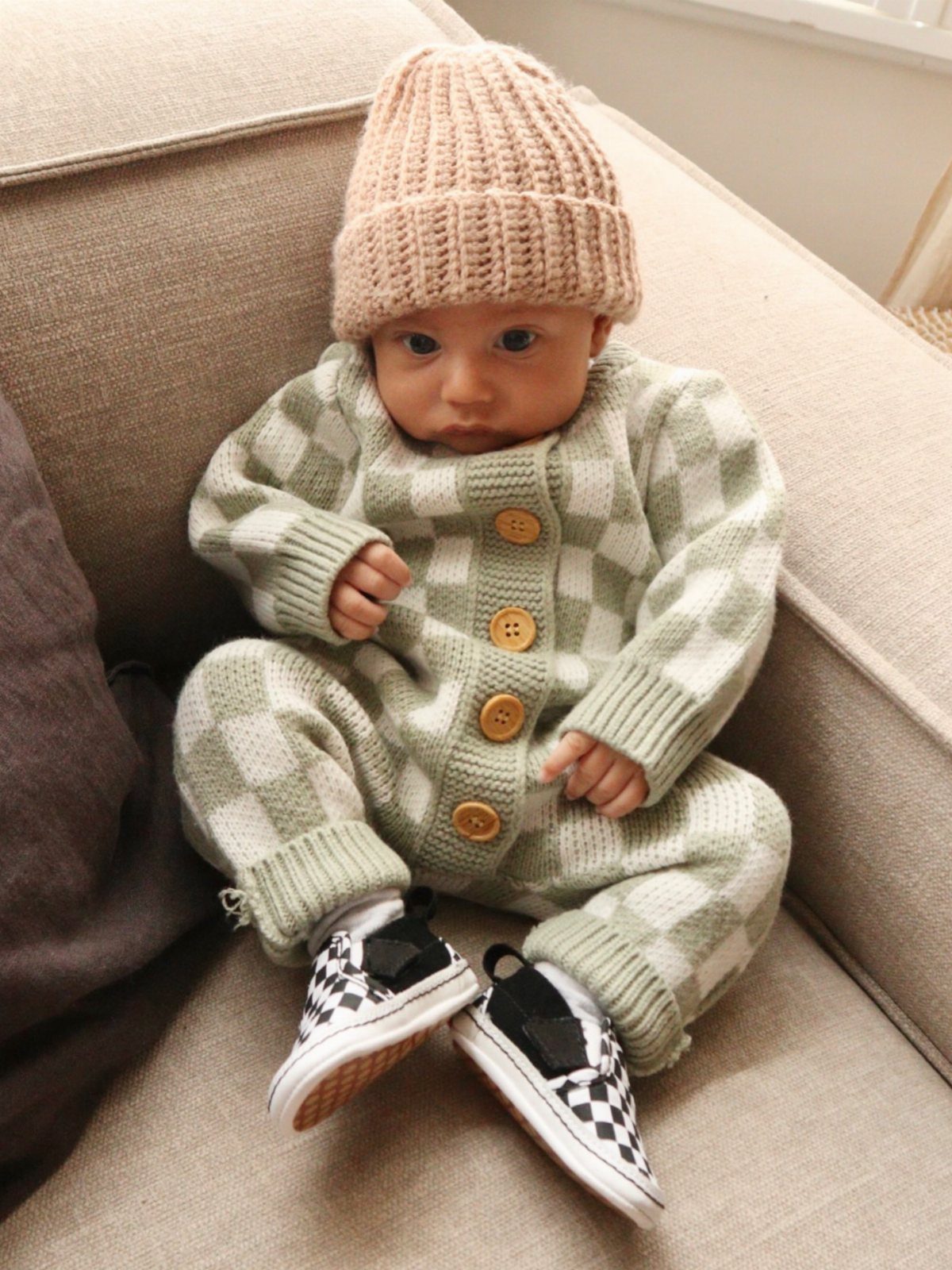 Baby wearing a green checkered knit romper, peach beanie, and checkered sneakers.
