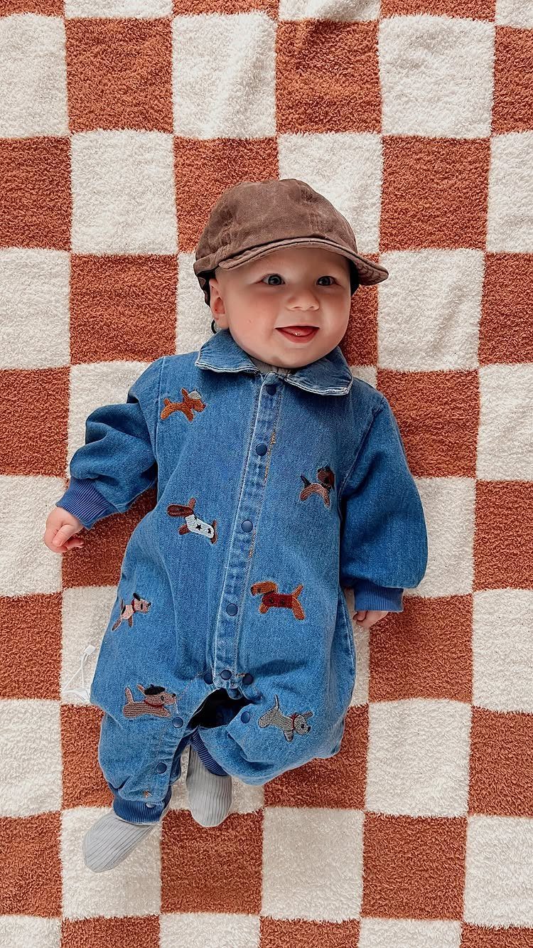 Baby in a blue denim jumpsuit with dog patches and a brown cap.