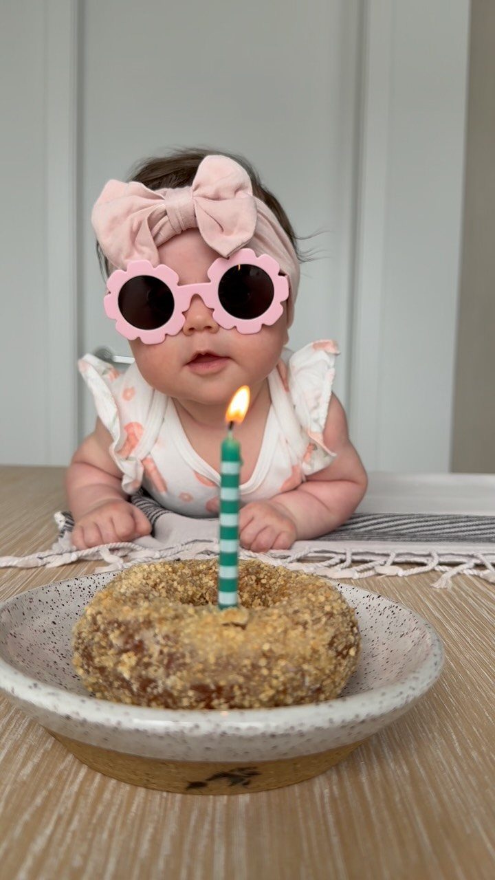 Baby wearing pink flower sunglasses and bow headband posing behind a birthday donut.