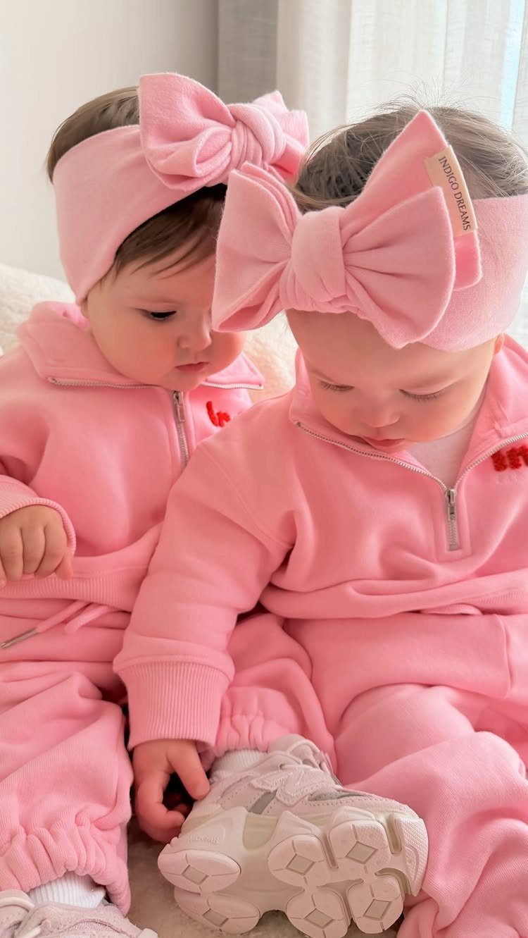 Two infants in matching pink zip-up sweatshirts, pants, and large bow headbands.