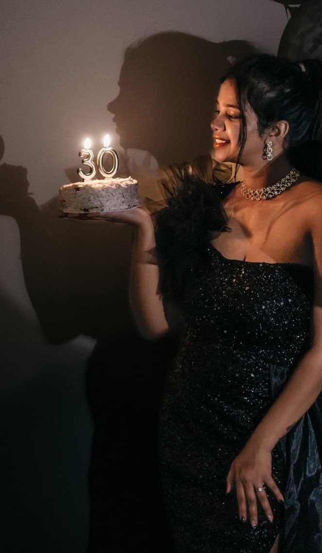 Woman in sparkling black dress holding a 30th birthday cake with lit candles.
