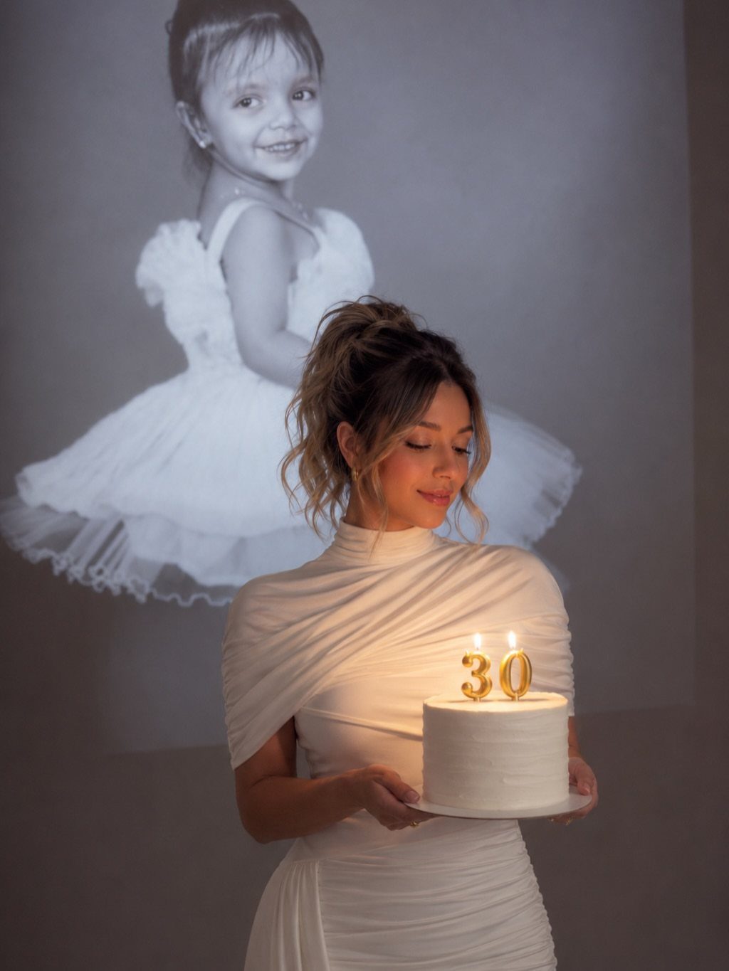 Woman in white draped dress holding 30th birthday cake with childhood photo projection.