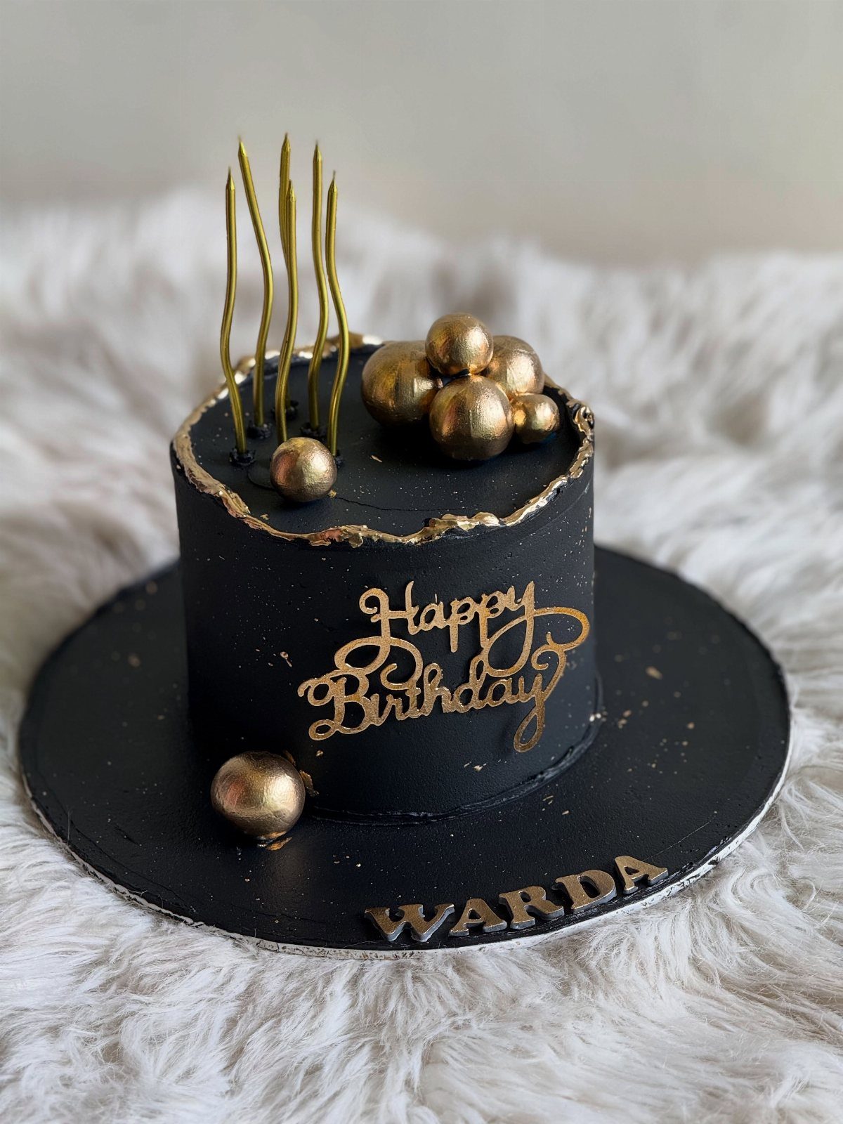 Black frosted birthday cake with gold spheres, gold candles, and gold lettering.