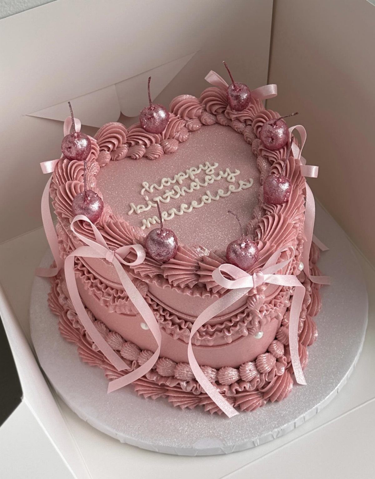 Heart-shaped pink birthday cake with glittery cherries, ribbons, and ornate frosting swirls.