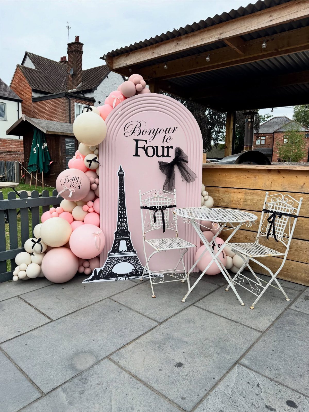 Parisian birthday setup with pink balloons, Eiffel Tower cutout, and white bistro furniture.