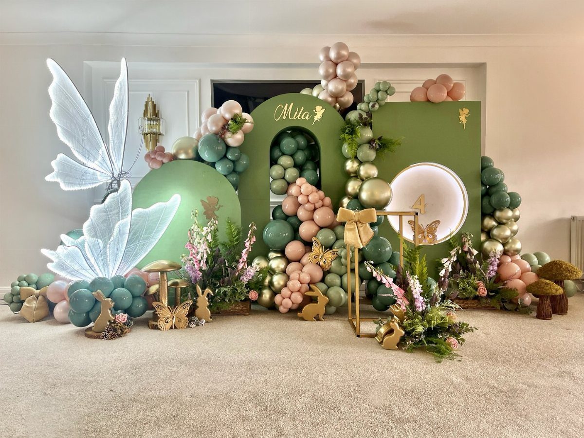 Green and gold balloon arch with floral arrangements and woodland decorations for a birthday.