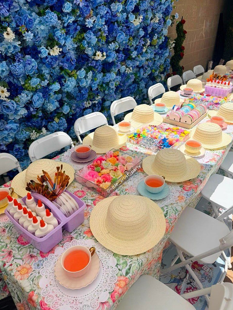 Tea party table setting with straw hats, tea cups, and blue flower wall.