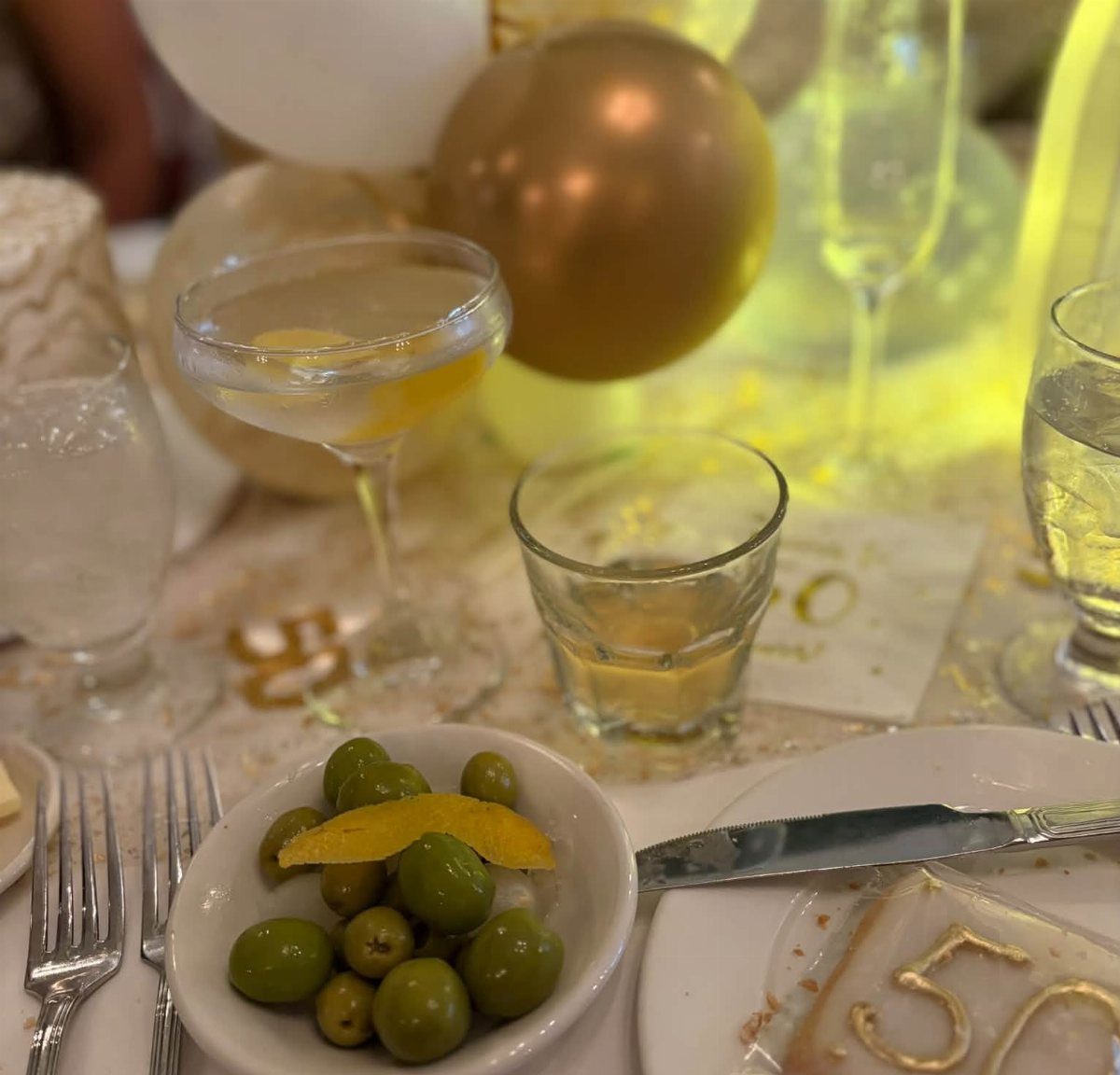 Party table setting with gold balloons, cocktails, olives, and gold number 50 decorations.