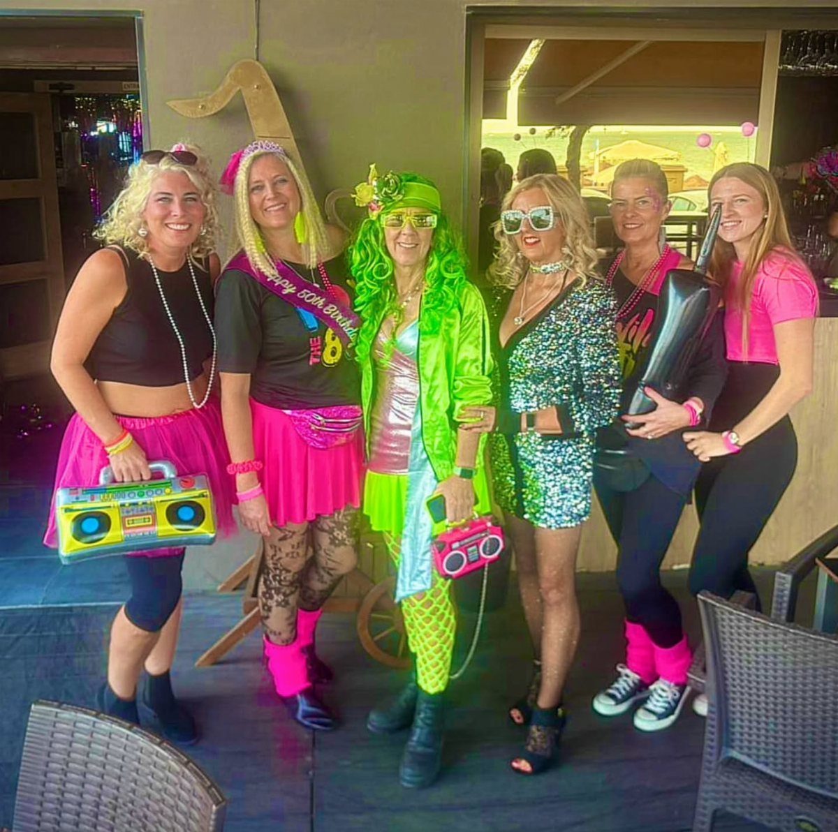 Six women in neon and sequined party outfits posing with a boombox outdoors.