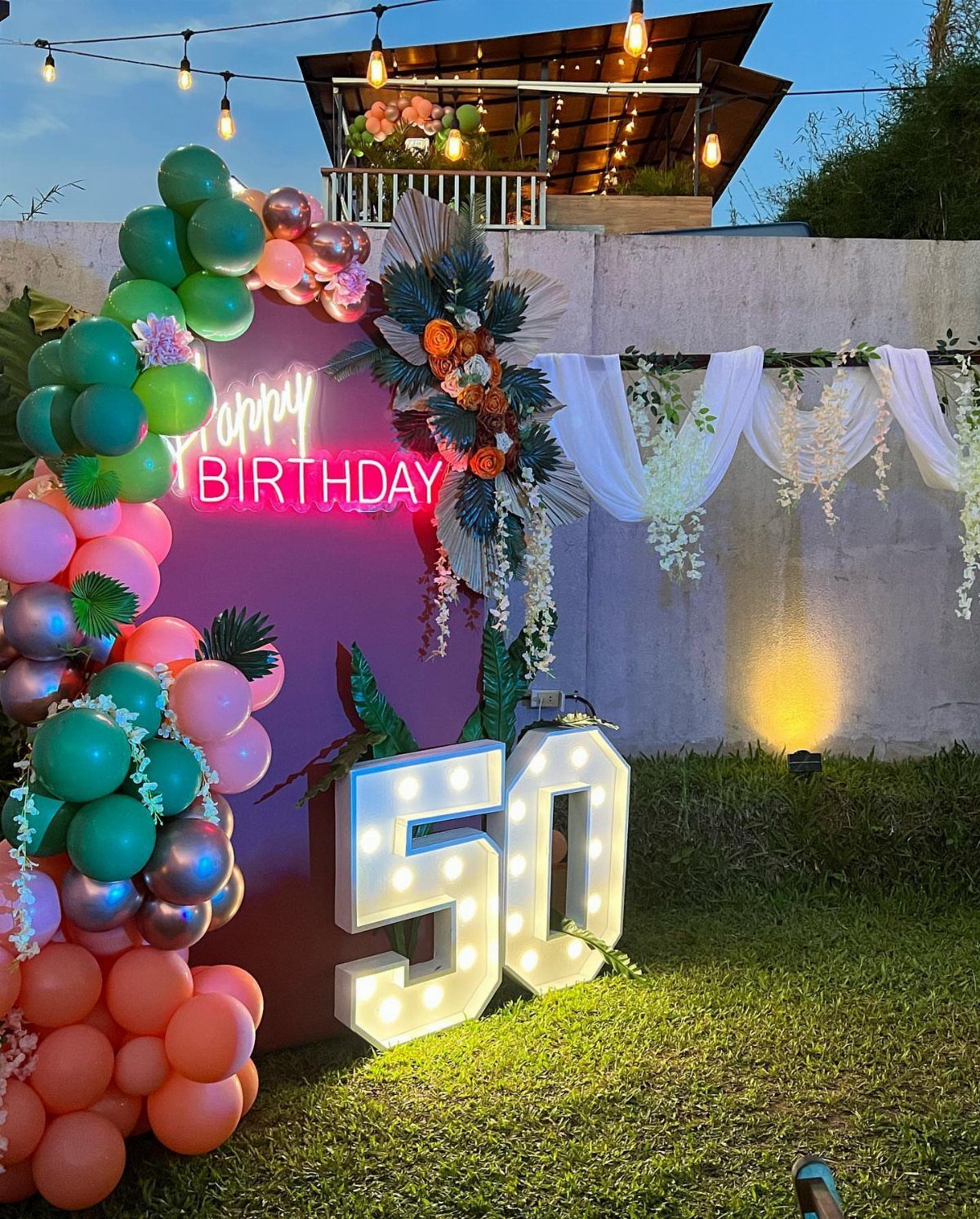 Illuminated number 50 sign with pink backdrop and colorful balloon arch outdoors.