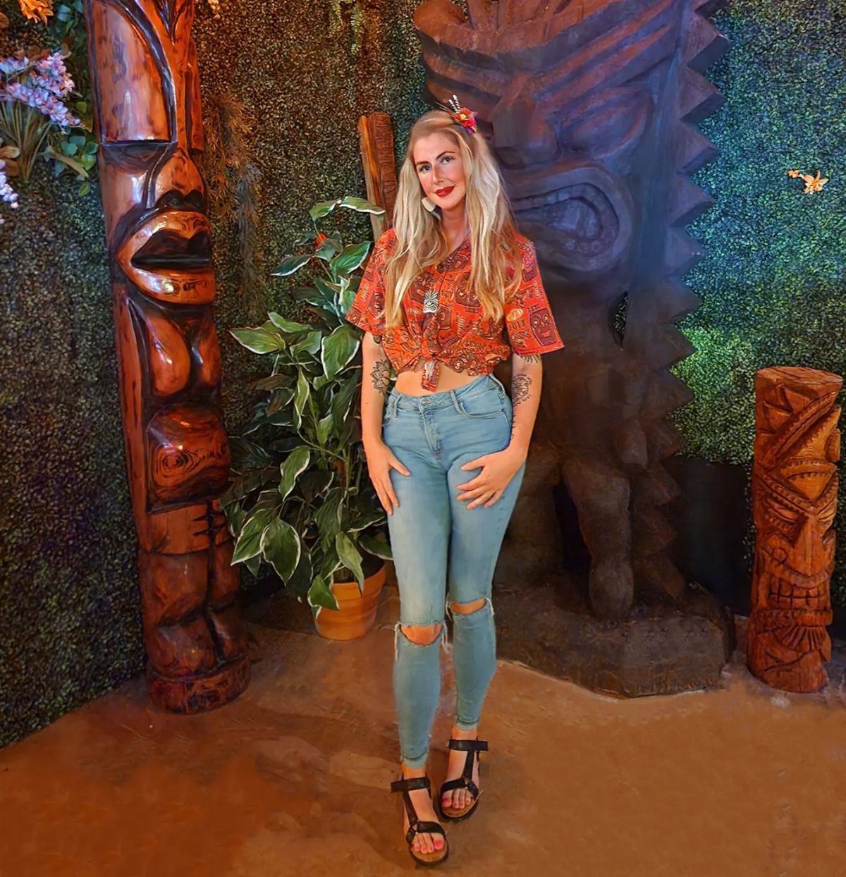 Woman in floral shirt posing with large carved wooden tiki totems and greenery.