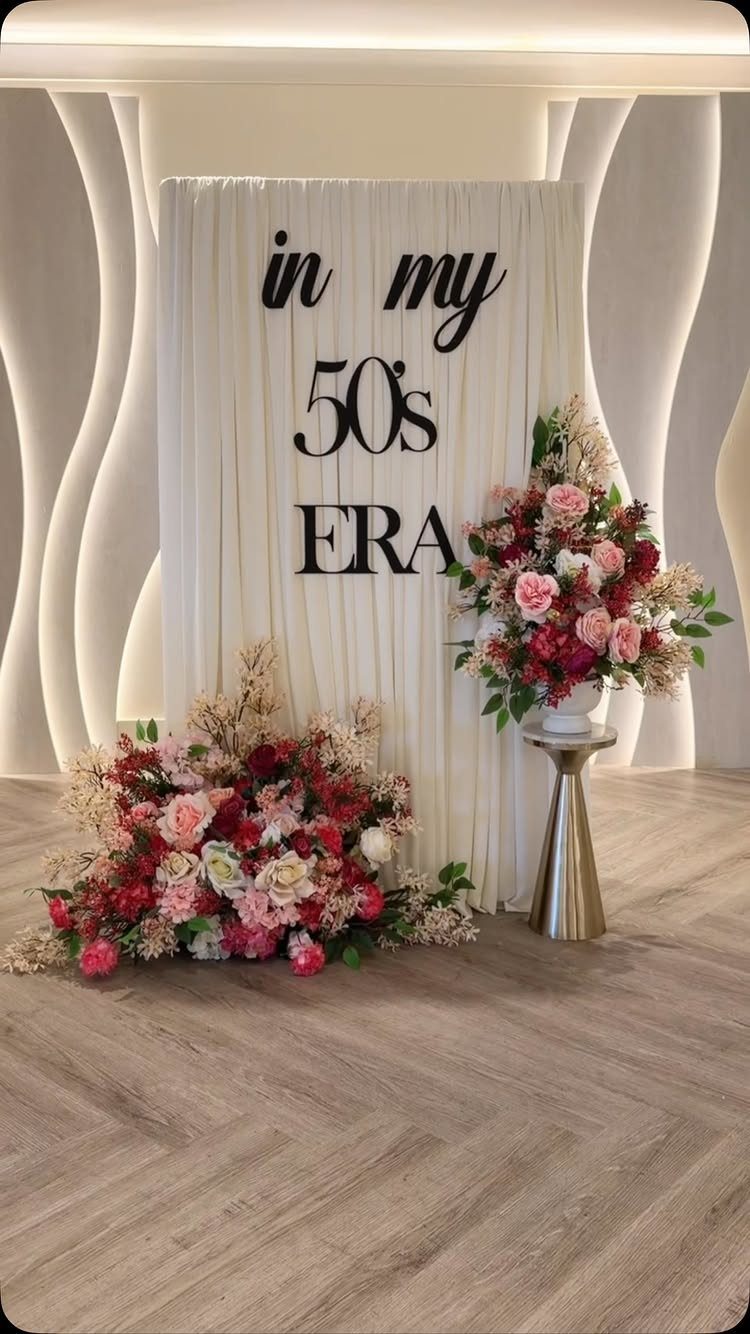 White fabric backdrop with 50s ERA text surrounded by pink and red floral arrangements