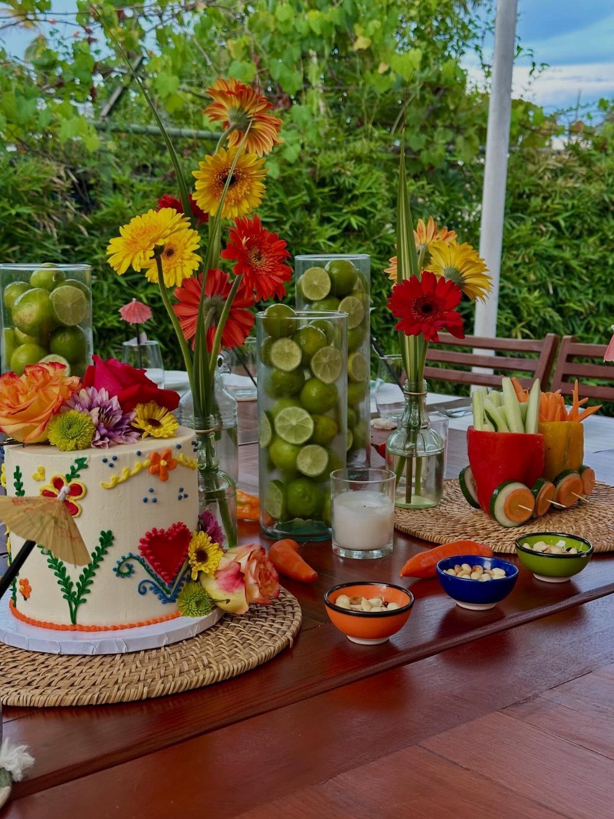 Festive outdoor table with a decorated cake, lime-filled vases, and colorful flowers.