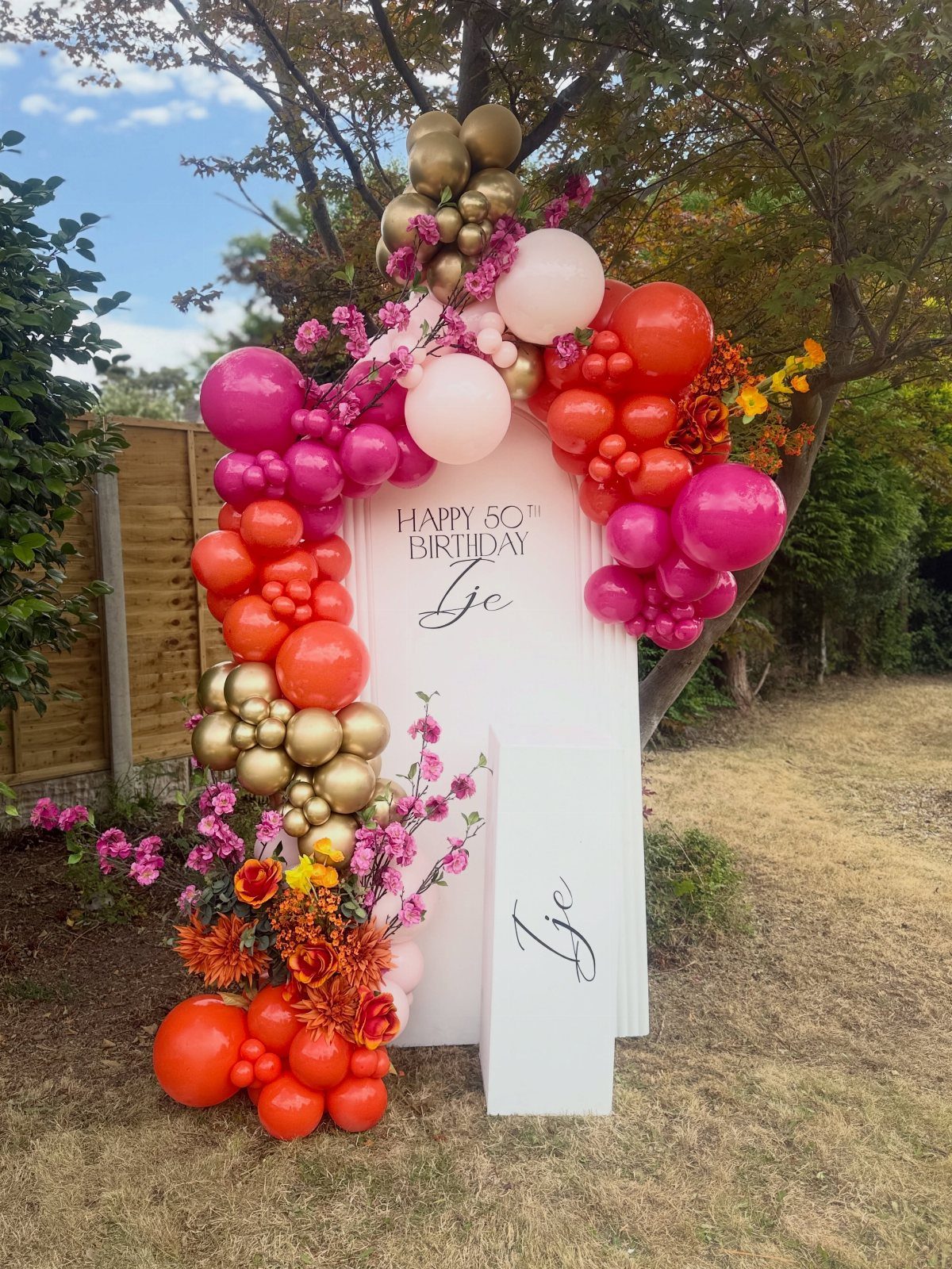 White arched birthday backdrop with pink, orange, and gold balloon garland and flowers.