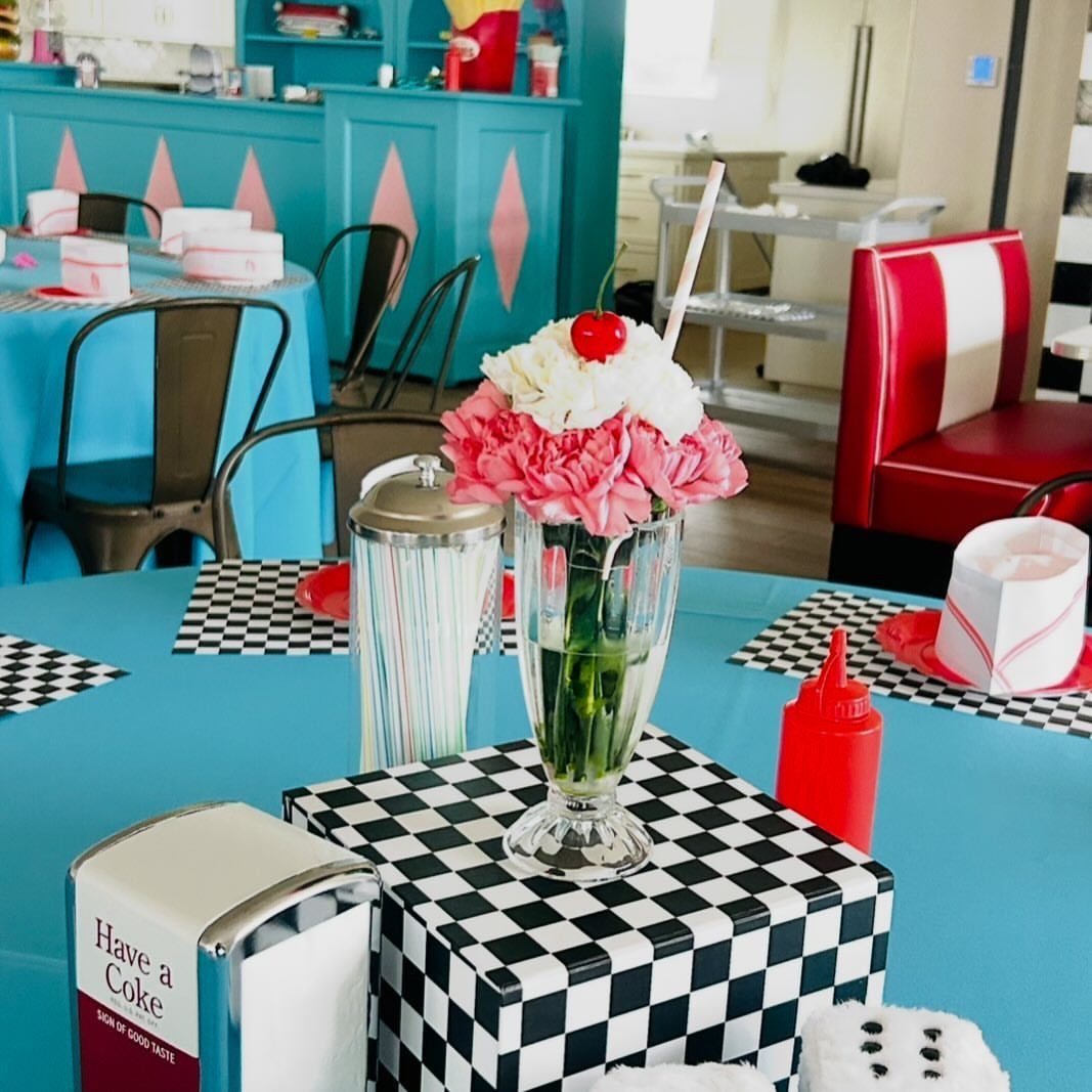 Diner-themed table setting with blue tablecloth, checkered patterns, and a floral milkshake glass.