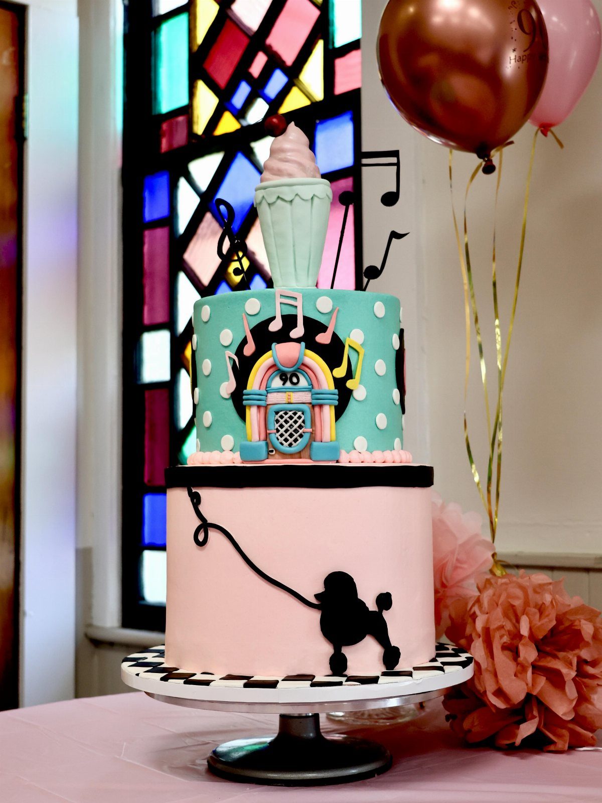 Multi-tiered 1950s themed cake with jukebox and poodle decorations on checkered stand.