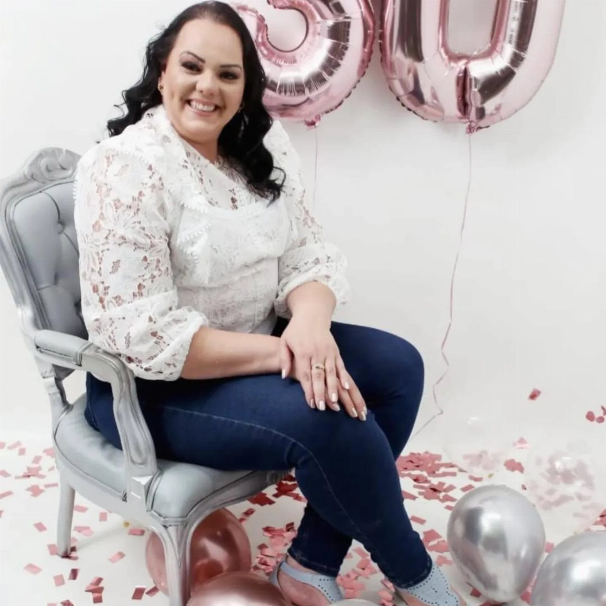 Woman wearing a white lace blouse and blue jeans sitting by birthday balloons.