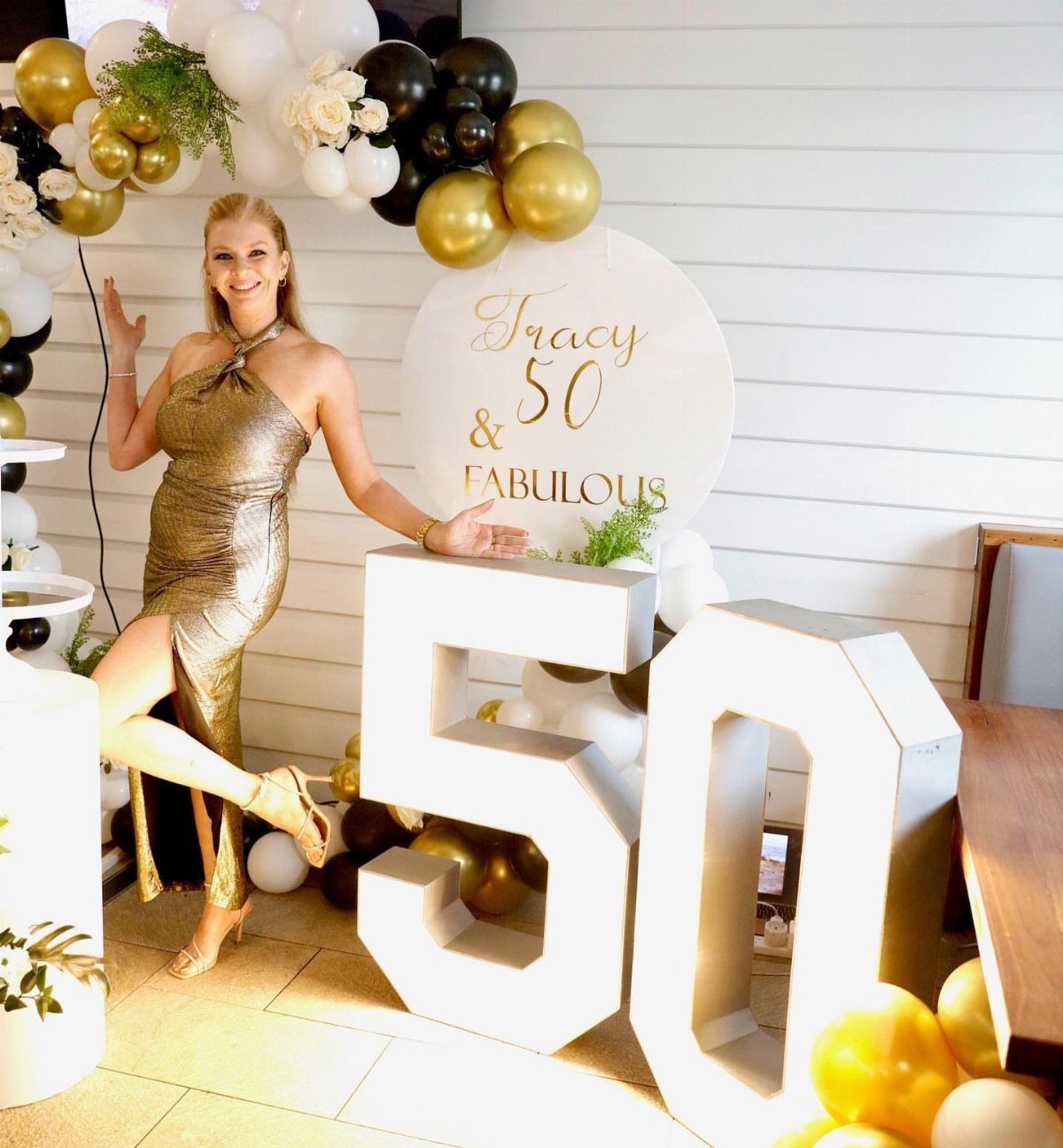 Woman in metallic gold dress posing by large white 50 numbers and balloons