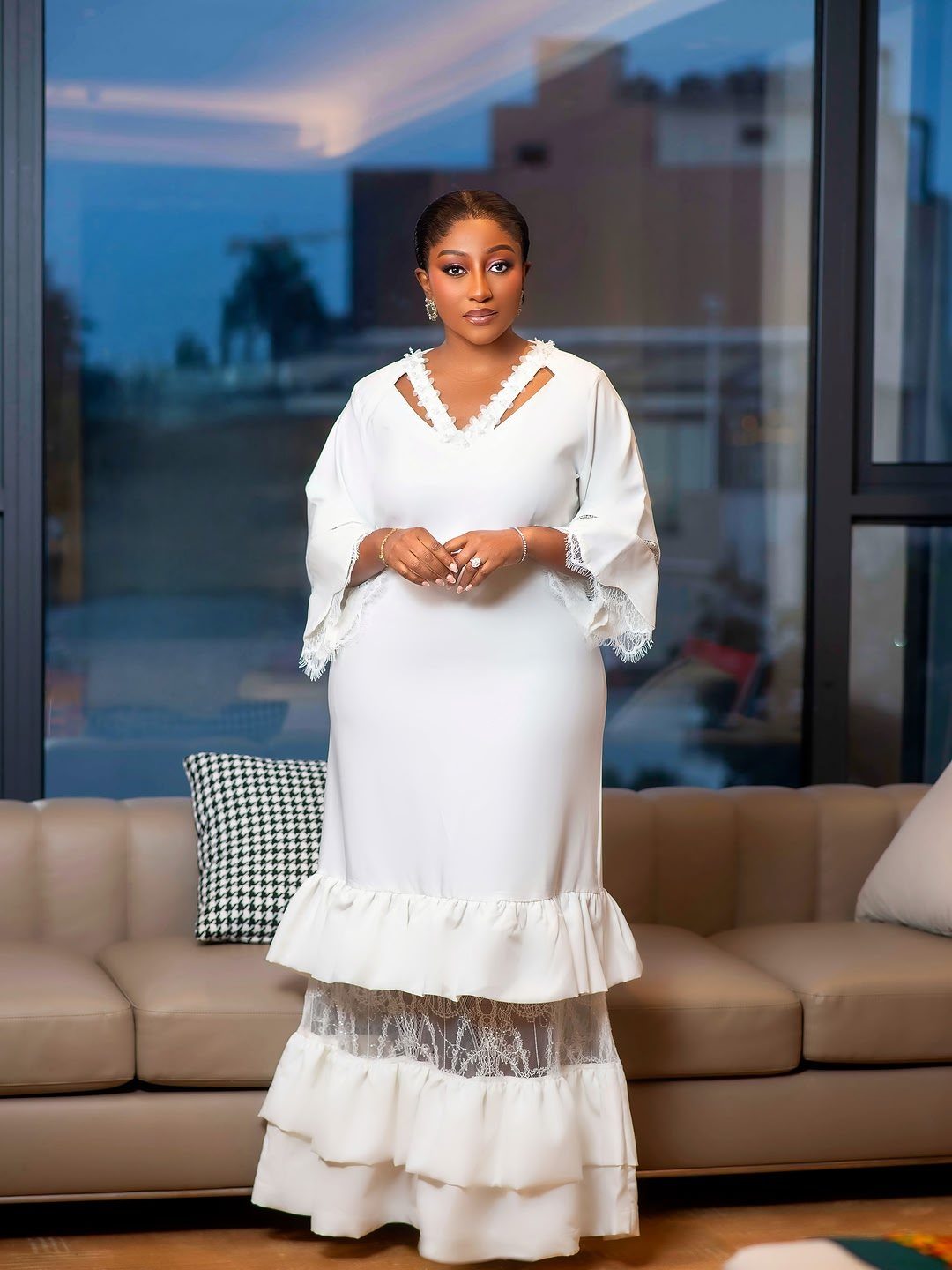 Woman in a long white tiered dress with lace details standing indoors.