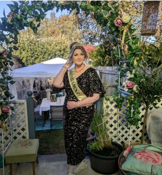 Mature woman in a black sequined dress and gold sash posing in a garden