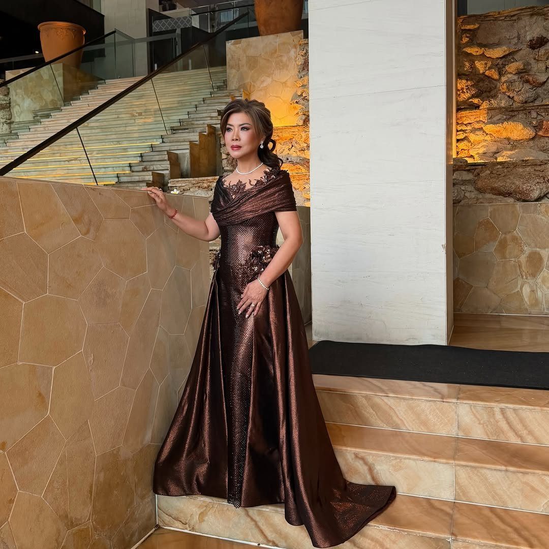 Woman in a shimmering bronze-brown floor-length gown posing by a stone wall.
