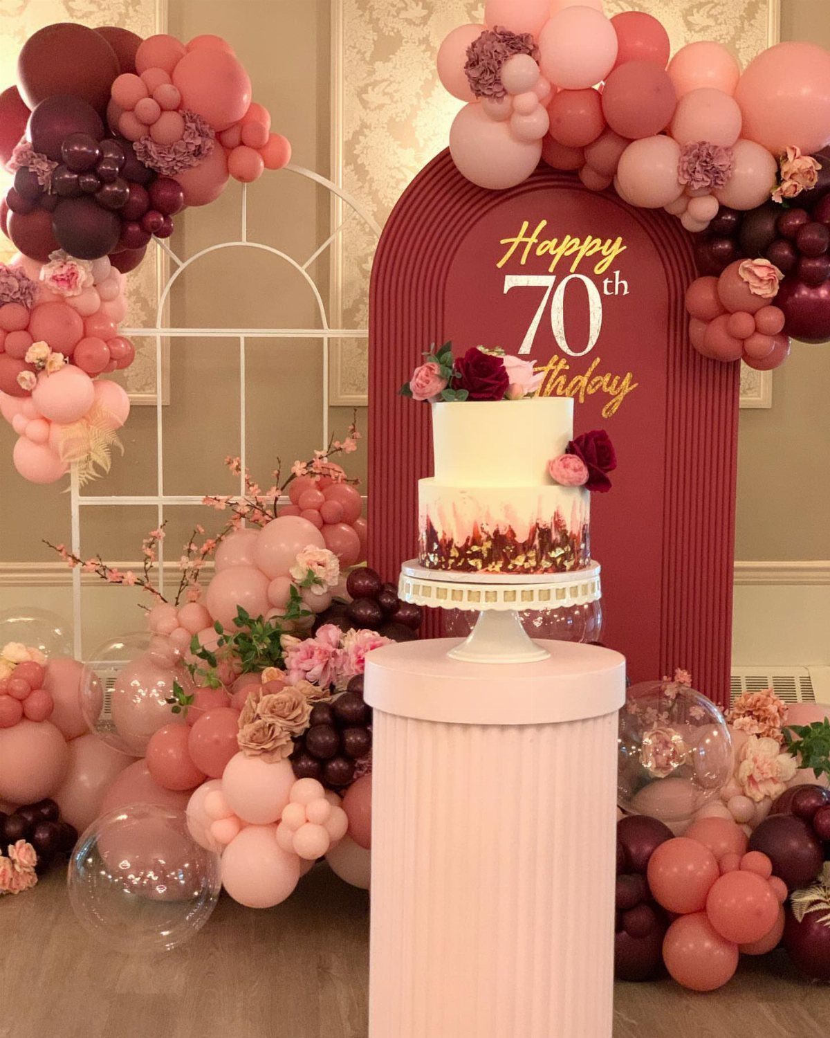 Two-tier birthday cake on a pink pedestal with pink and burgundy balloon decorations.