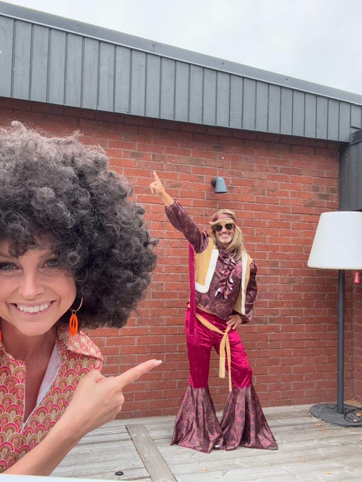 Two people wearing 1970s disco costumes posing outdoors against a brick wall.