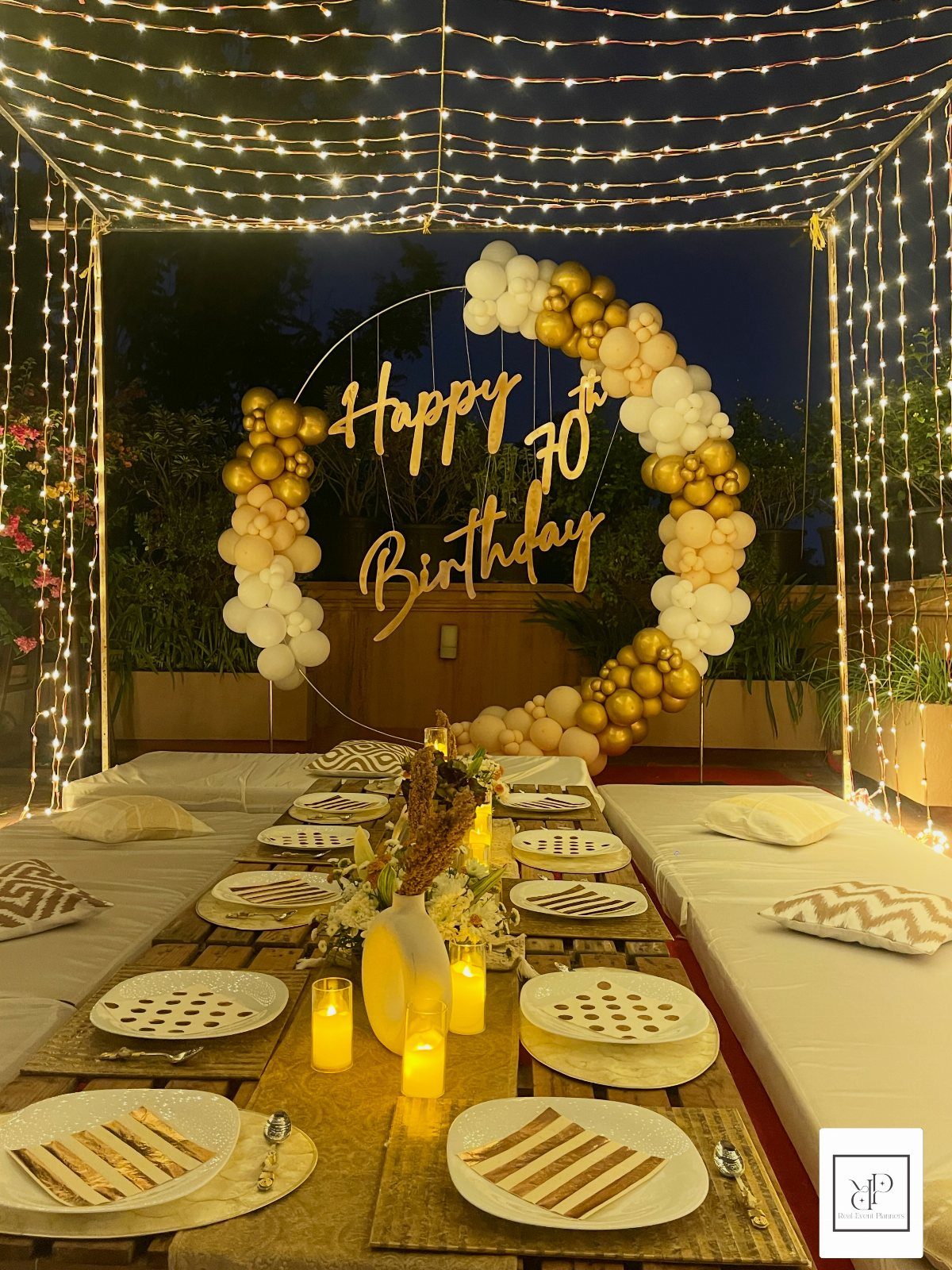 Outdoor 70th birthday dinner table with gold balloon arch and fairy light canopy.