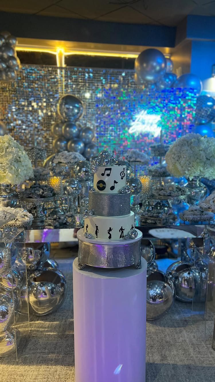 Three-tier disco themed cake with musical notes before a silver sequin backdrop.