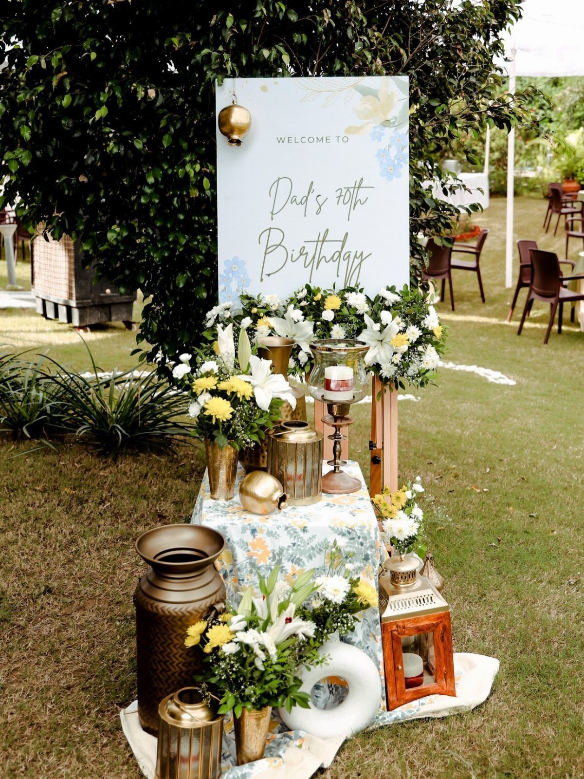 Outdoor 70th birthday welcome sign with floral arrangements and gold lanterns on grass