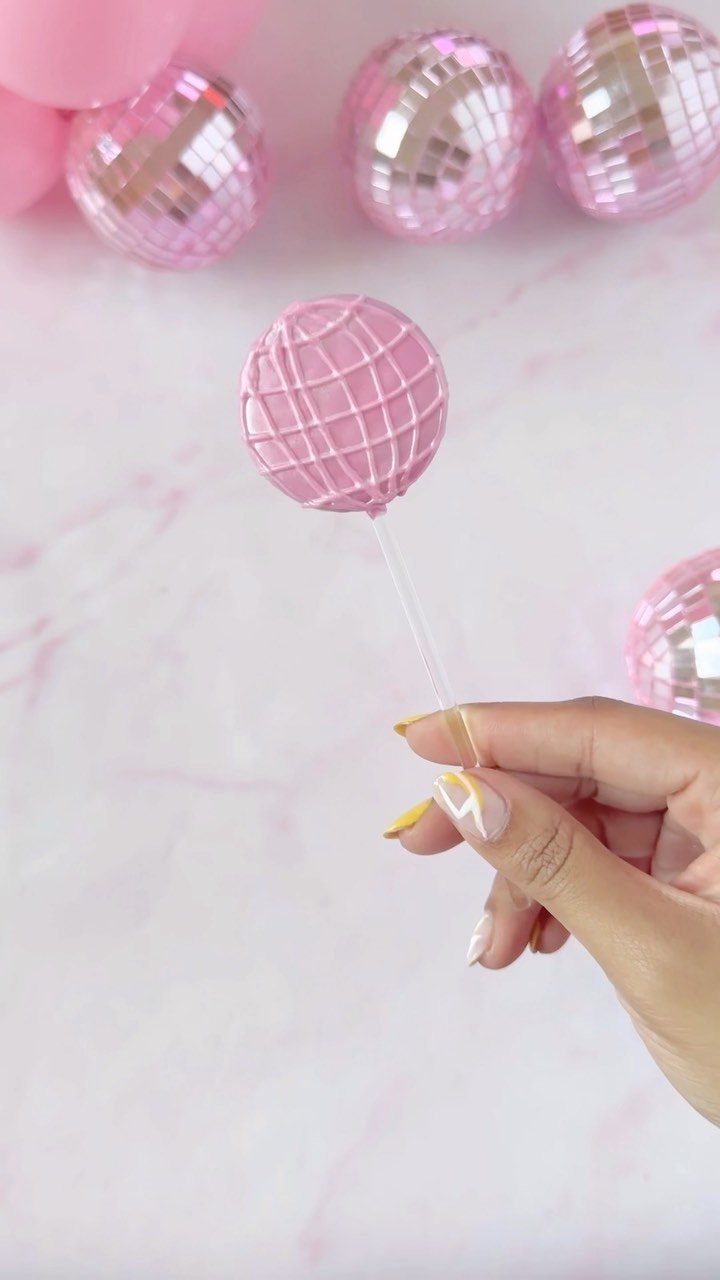 Pink disco ball cake pop held by a hand against a marble background.