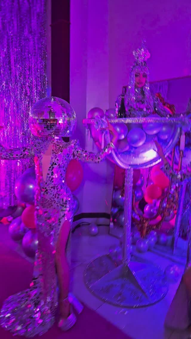 Person in sequined dress and disco ball mask next to balloon display