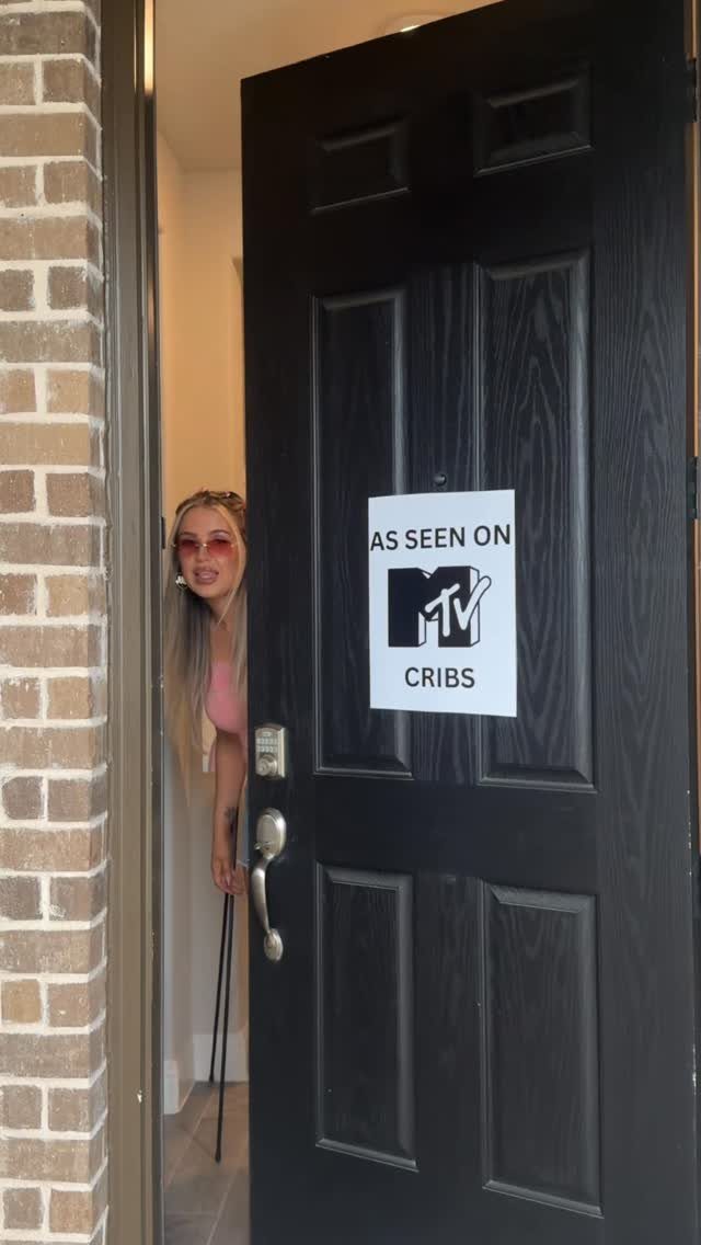 Woman peeking behind a black door with an MTV Cribs sign attached.