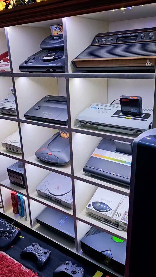 Vintage video game consoles and controllers displayed on white shelving units.