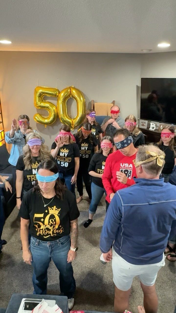 Adults wearing blindfolds and birthday shirts standing in a room with gold balloons.