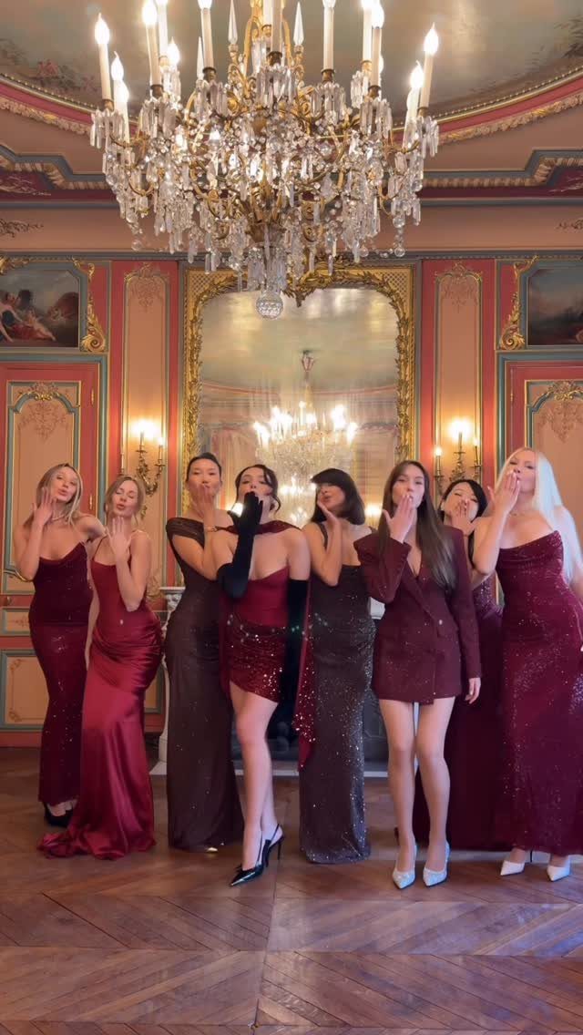 Women in red formal attire posing under a crystal chandelier in an ornate room.