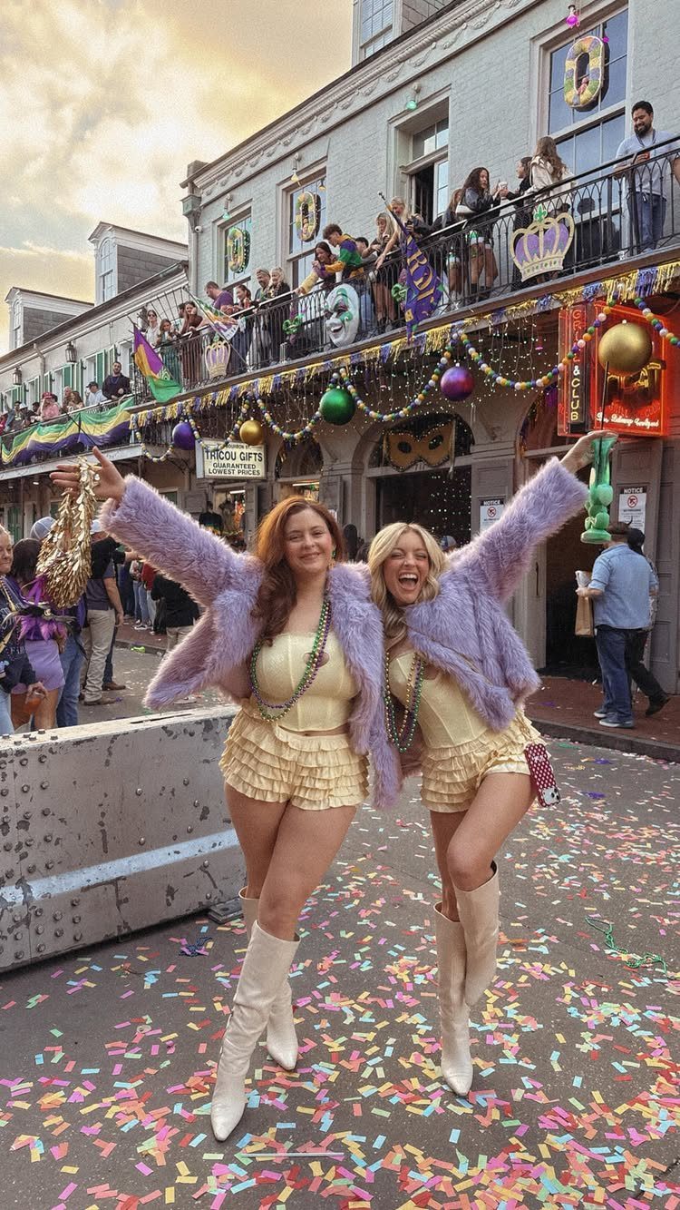 Two women in purple coats and yellow outfits celebrating on a confetti-covered street.